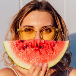 Charming lovely woman in yellow sunglasses hiding a half of her face with piece of watermelon, joking, standing against the wall, outdoors.